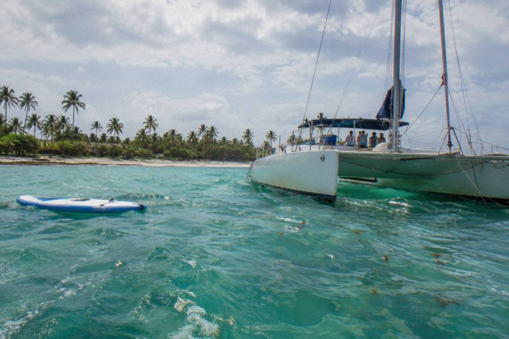 Catamaran anchored in turquoise water with a paddleboard floating beside it and guests seated on deck