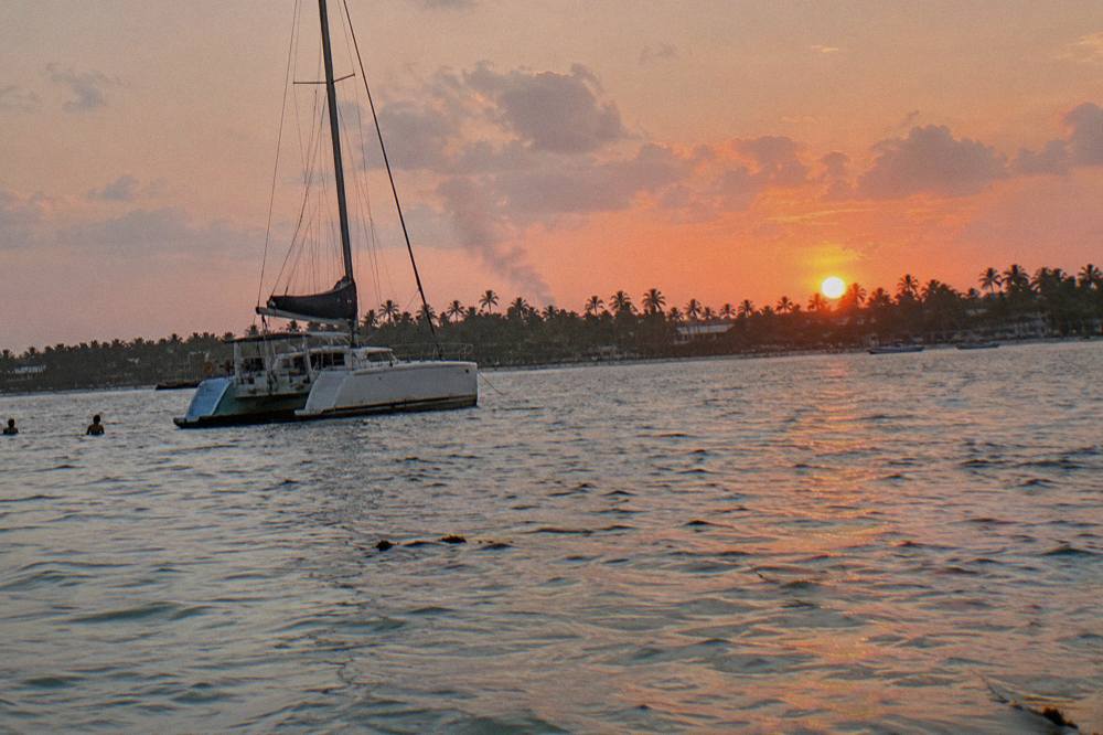 A catamaran at anchor on calm water under a wide pink and orange evening sky with a dark palm shoreline in the distance