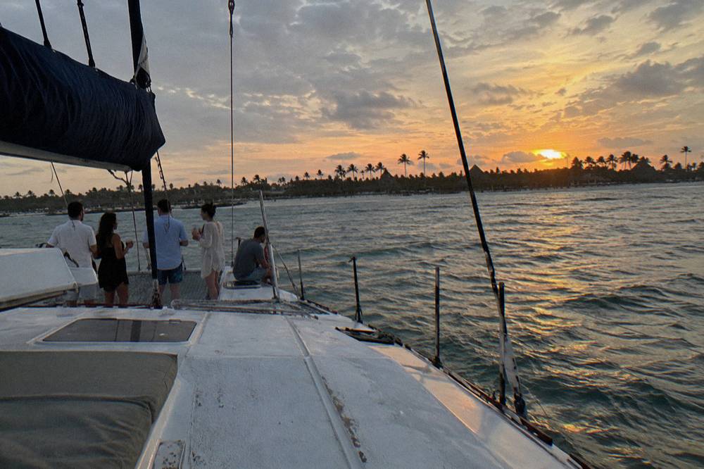 Guests seated along the deck rail of a catamaran with drinks as the sky glows orange over calm water