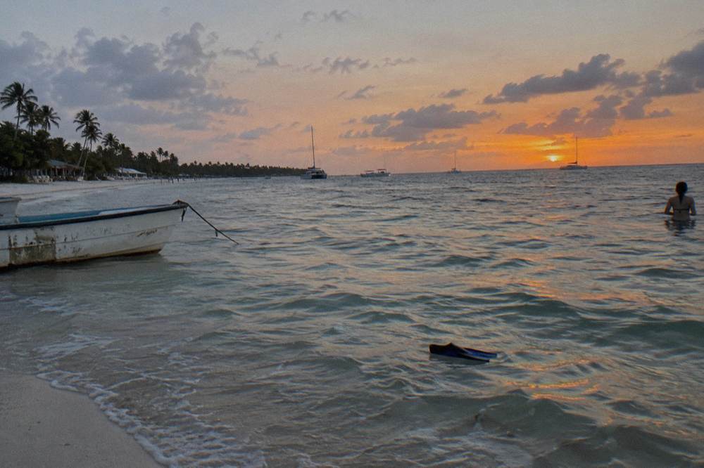 Shallow sandbar water mirroring a violet and orange sky with distant boat silhouettes