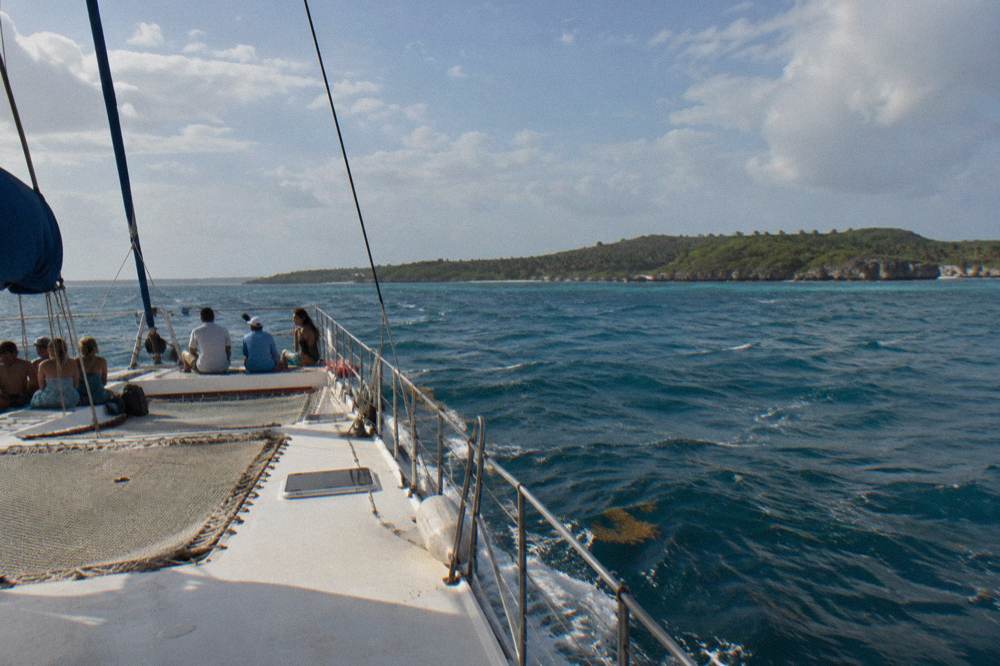 Passengers on a catamaran deck in open water with green island coastline on the horizon