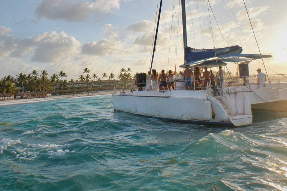 A wide party catamaran crowded with standing guests dancing near a bar area