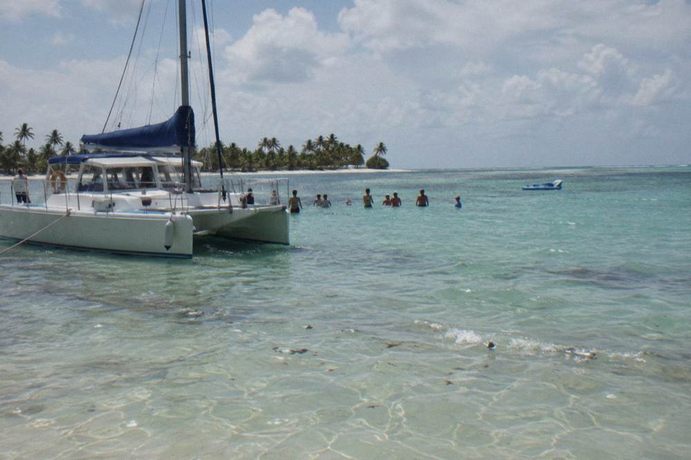 Guests in waist-deep water at a natural sandbar pool beside a moored catamaran