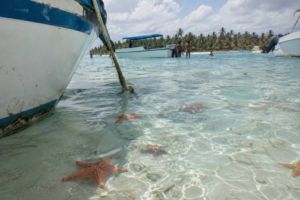 Shallow natural swimming pool with starfish visible on the sandy bottom beside anchored boats