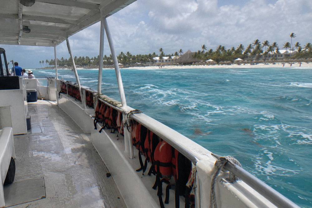 Covered tour boat side deck with orange life jackets on the rail, a cooler, and Bavaro beach beyond
