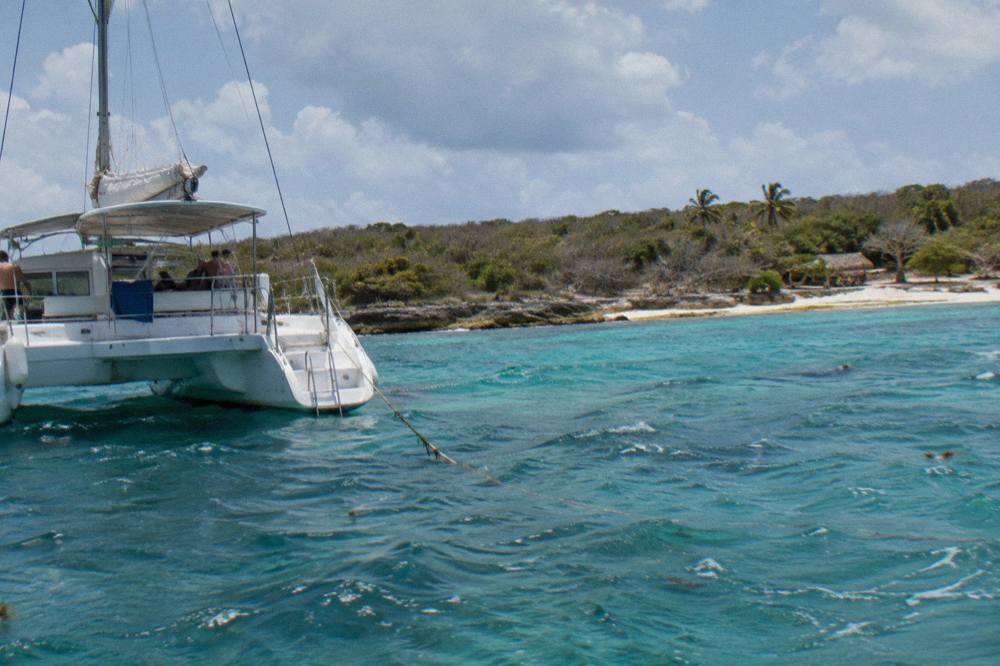 A white catamaran anchored in clear shallow water beside a sandy island beach