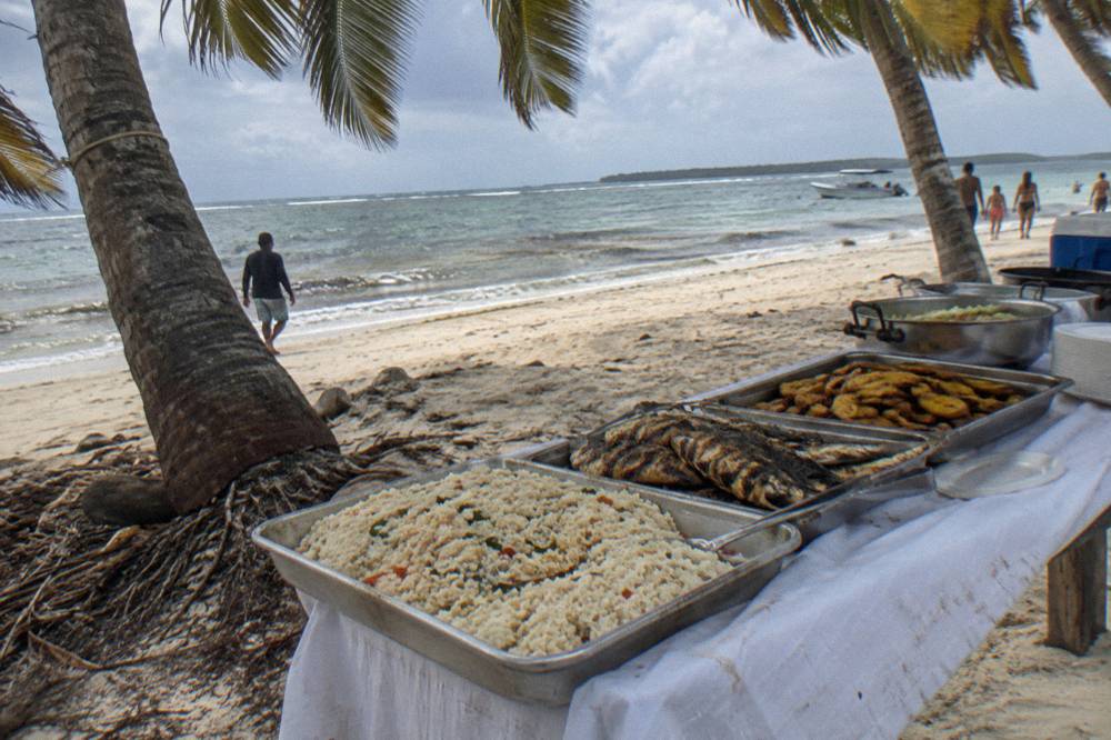 A buffet lunch table set on white sand under palm trees beside calm sea