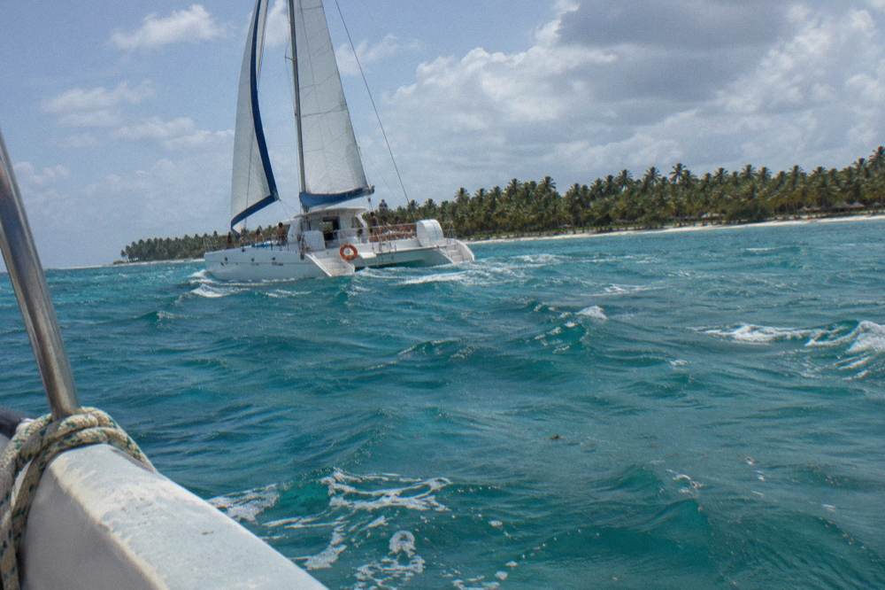 A sailing catamaran with guests on deck in open turquoise water near an island