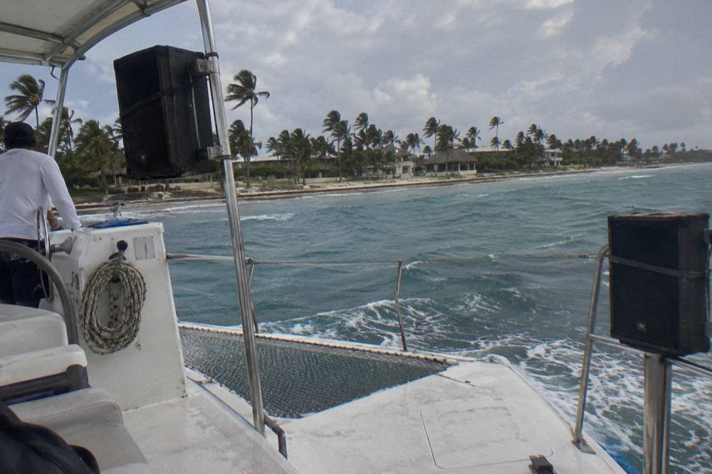 Open-air catamaran cruising along a calm turquoise coastline with music equipment on deck