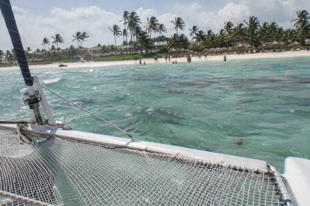 Empty catamaran bow netting over shallow green water with a beach and swimmers in the far distance