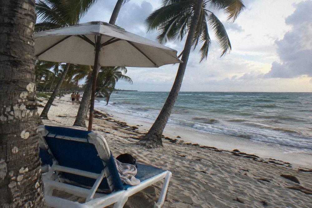 Beach bag, towel and sandals on sand beside shaded loungers under palms
