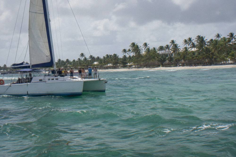 A catamaran under sail seen from the water with a handful of passengers standing at the bow rail