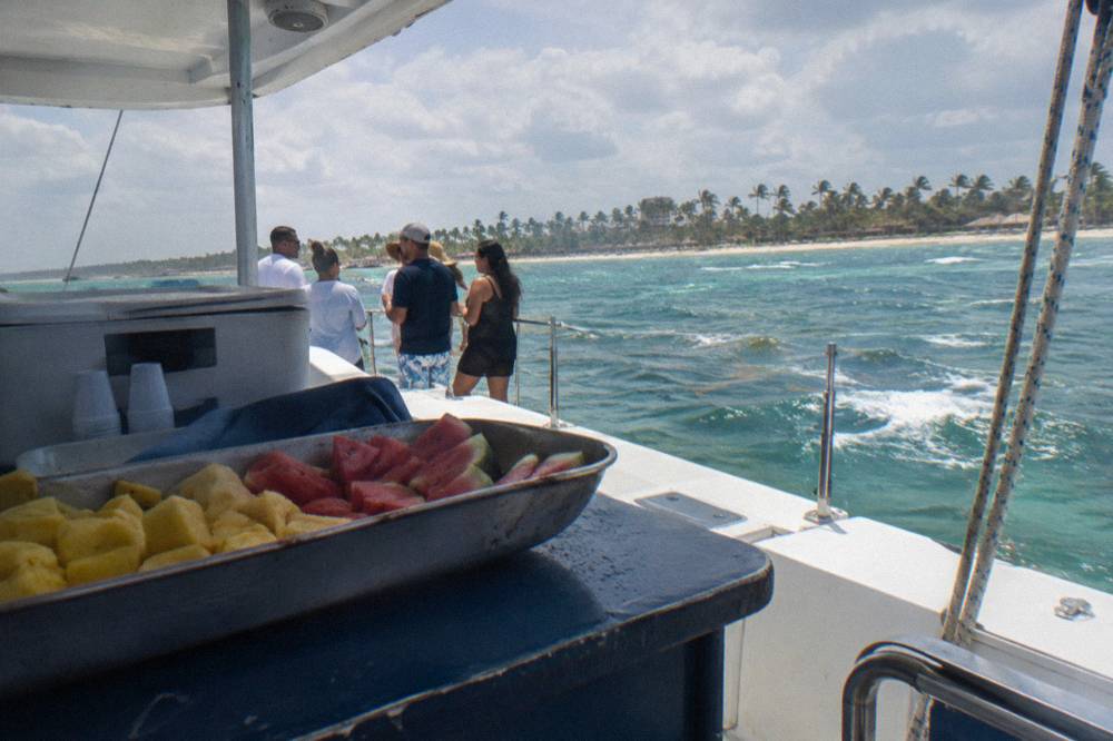 Tray of sliced pineapple and watermelon with plastic cups on a deck counter, guests at rail behind