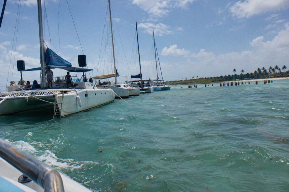 Several catamarans anchored in a line beside a shallow sandbar under a blue sky