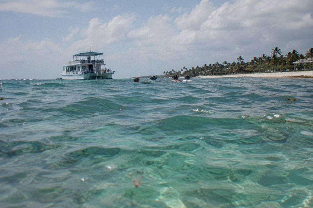 Double-deck tour boat anchored off a palm beach seen from water level with two or three distant swimmers