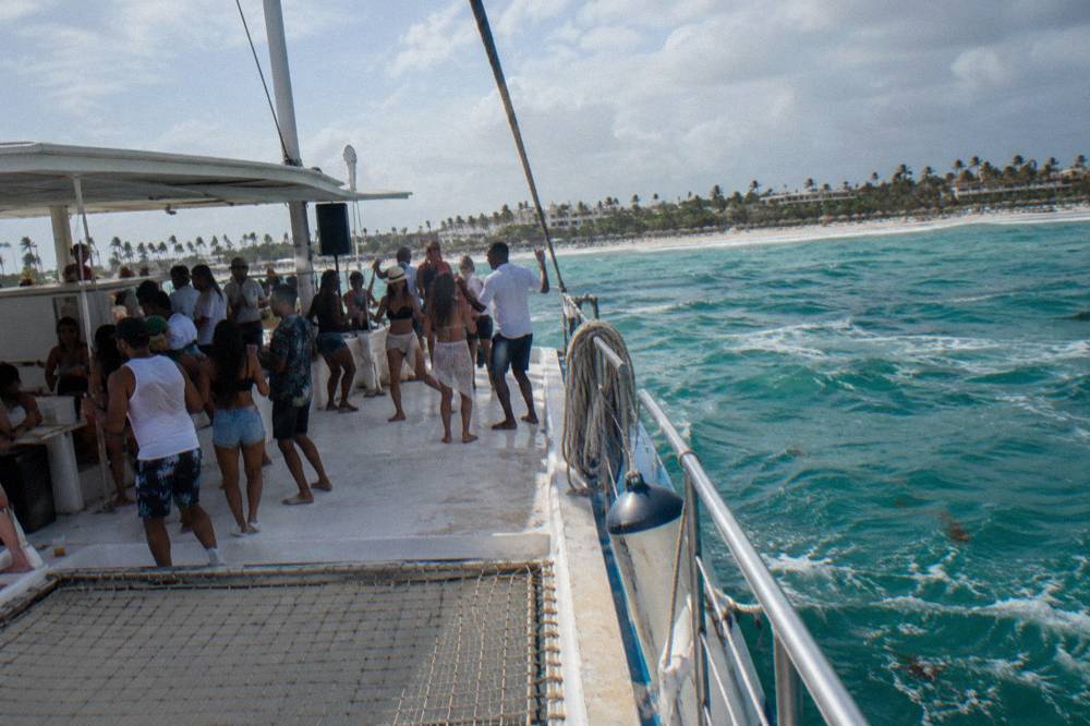 Crowded shared catamaran deck with guests dancing near a DJ booth under an open sky