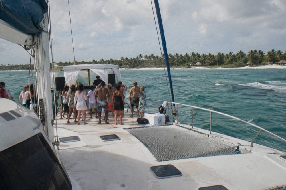 A catamaran deck crowded with guests dancing while a DJ booth plays near the bow