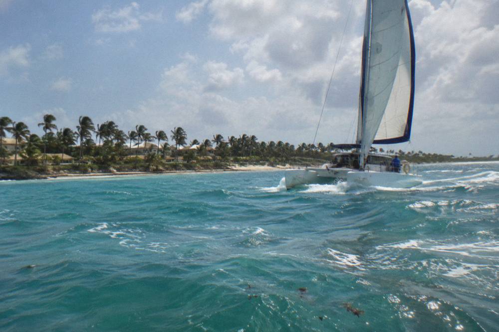 A catamaran under sail on calm open water with scattered clouds on the horizon