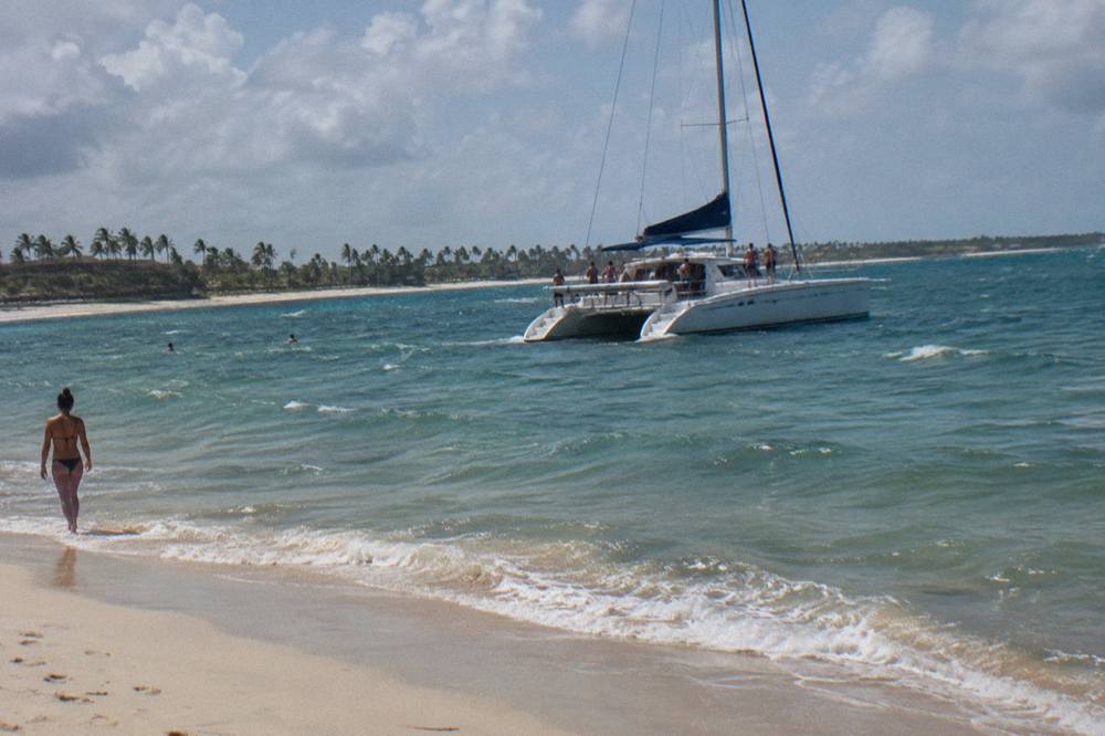 A shared catamaran with passengers dancing on deck near a sandy beach