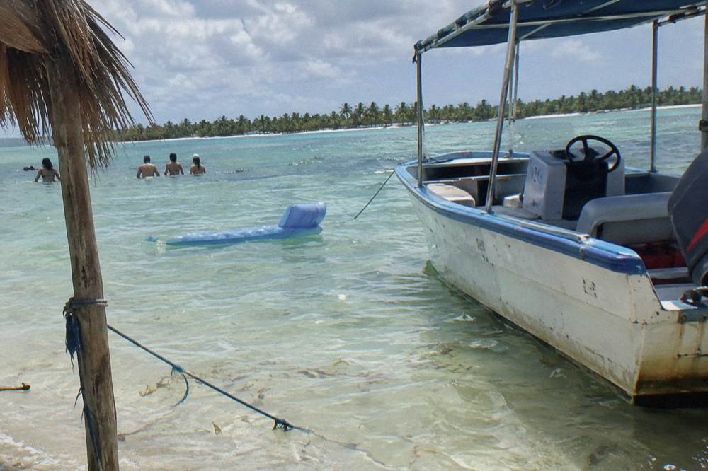 A shallow sandbank with people standing in clear water beside a moored boat