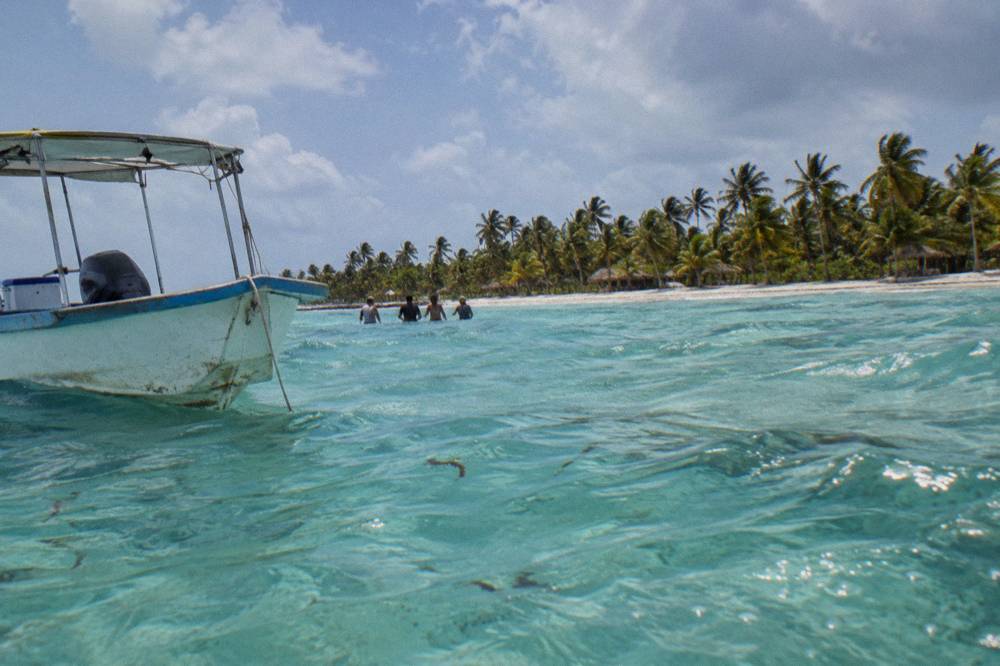 A shallow sandbar with people standing in waist-deep clear water beside a moored boat