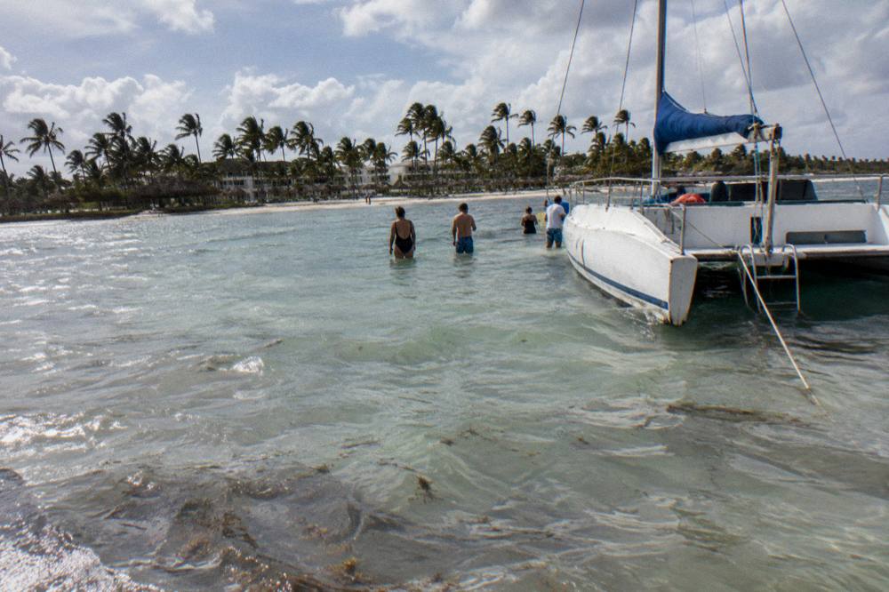 Small catamaran anchored at a shallow sandbar with guests standing in clear water