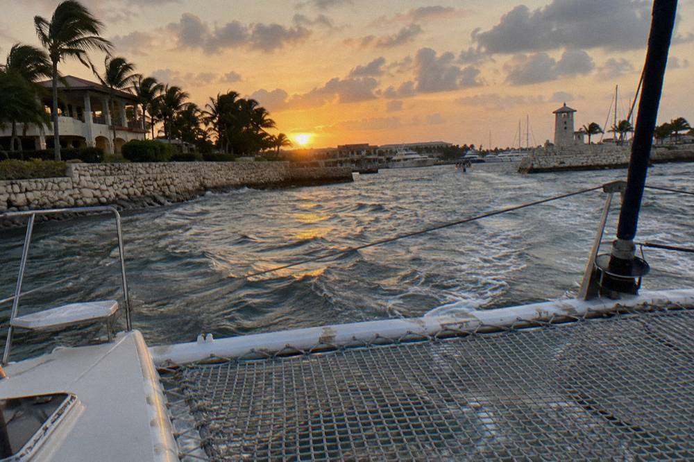 Open deck of a catamaran with nets and railings gliding over calm water as the sky turns orange near the coast
