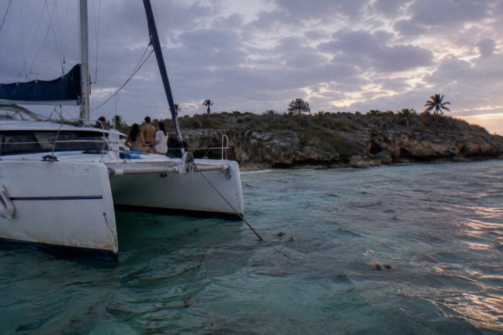 Private catamaran anchored in shallow clear water with a small group gathered at the bow under a soft pink sky
